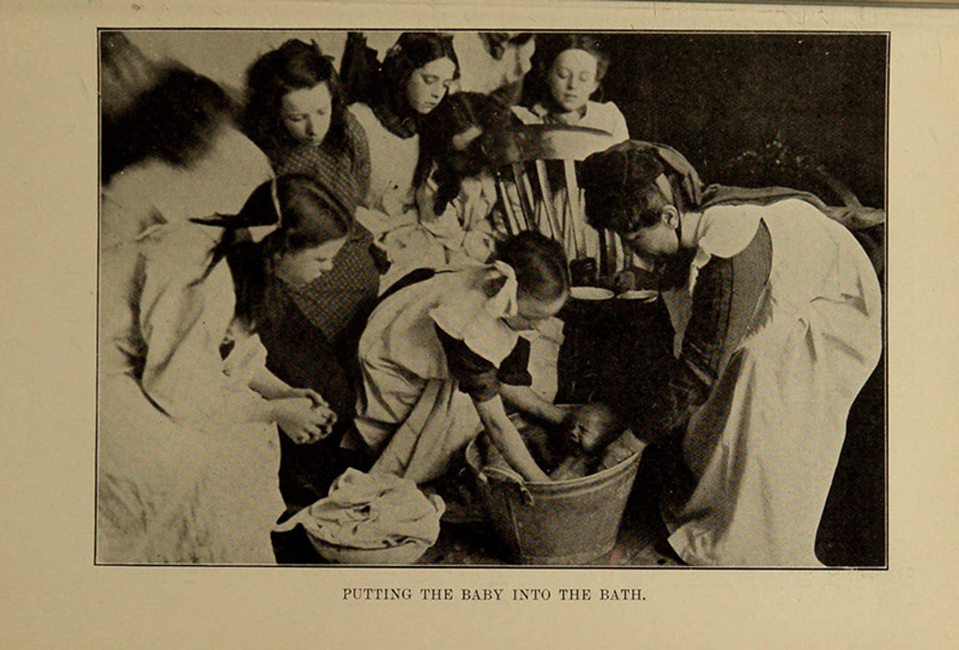 Vintage photo of women gathered around a tub bathing a baby, captioned “Putting the Baby into the Bath”.