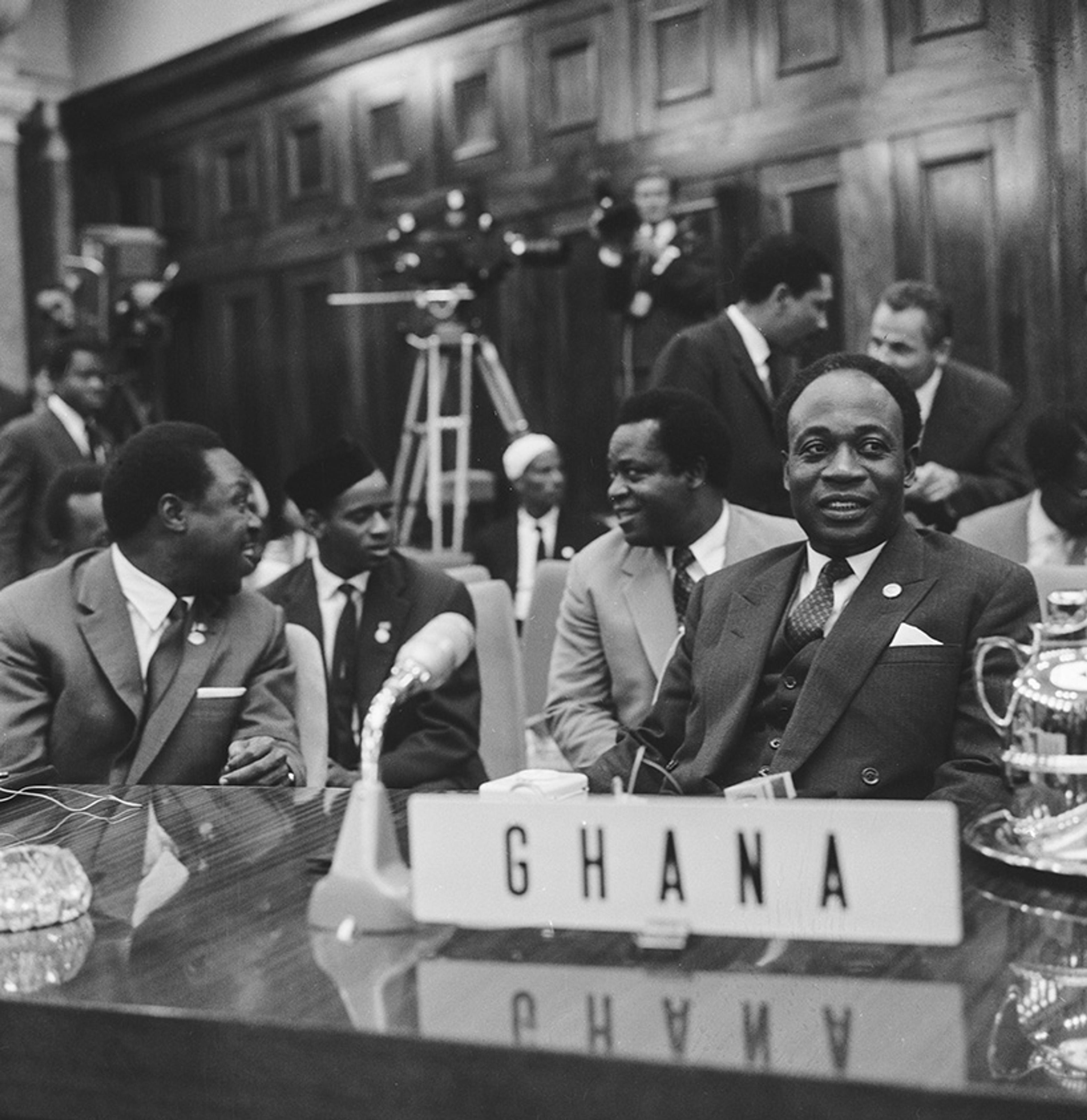 Vintage black and white photo of men wearing suits sitting at a conference table with a “GHANA” sign