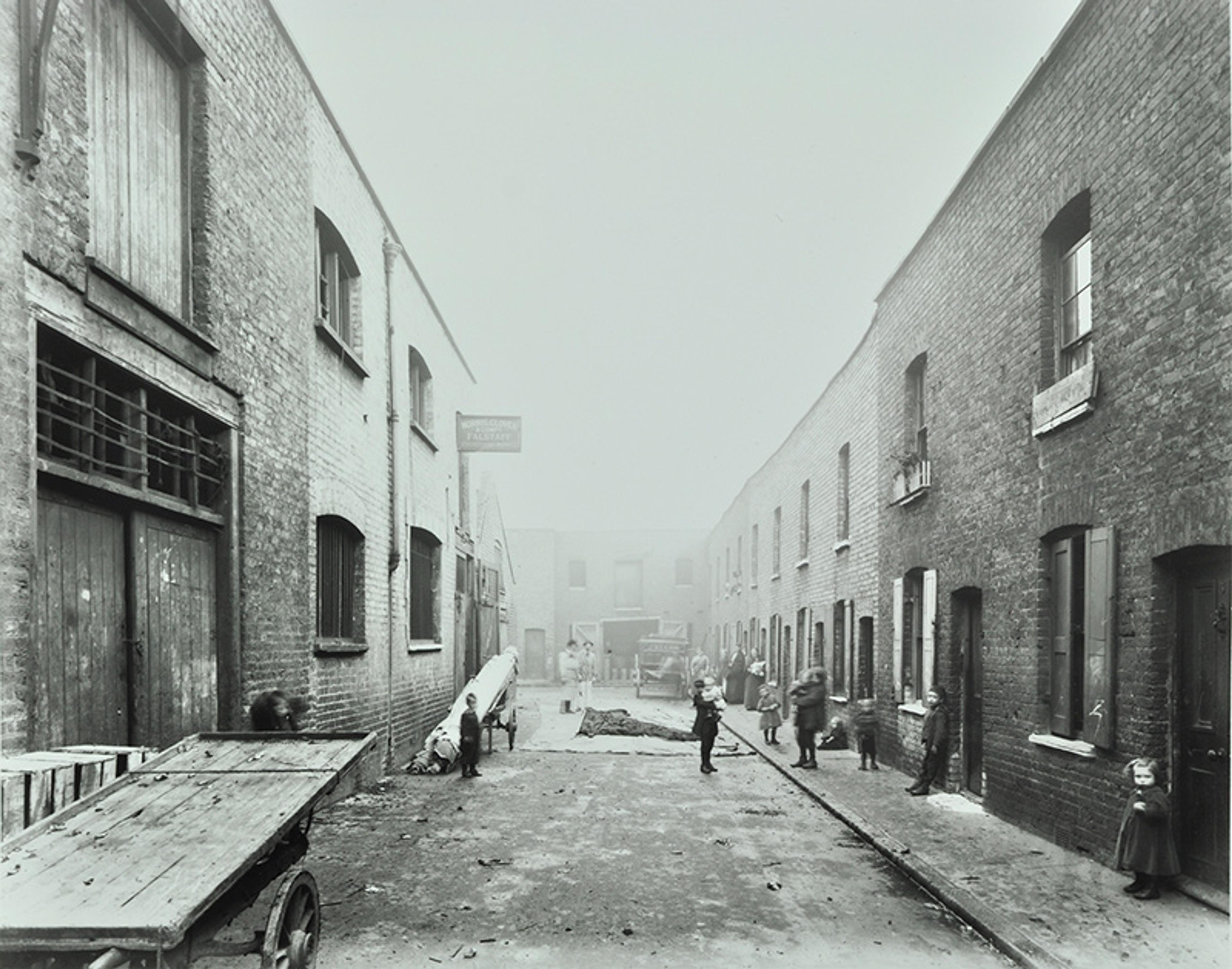 Vintage black and white photo of a narrow street with Victorian buildings, scattered people and children playing.