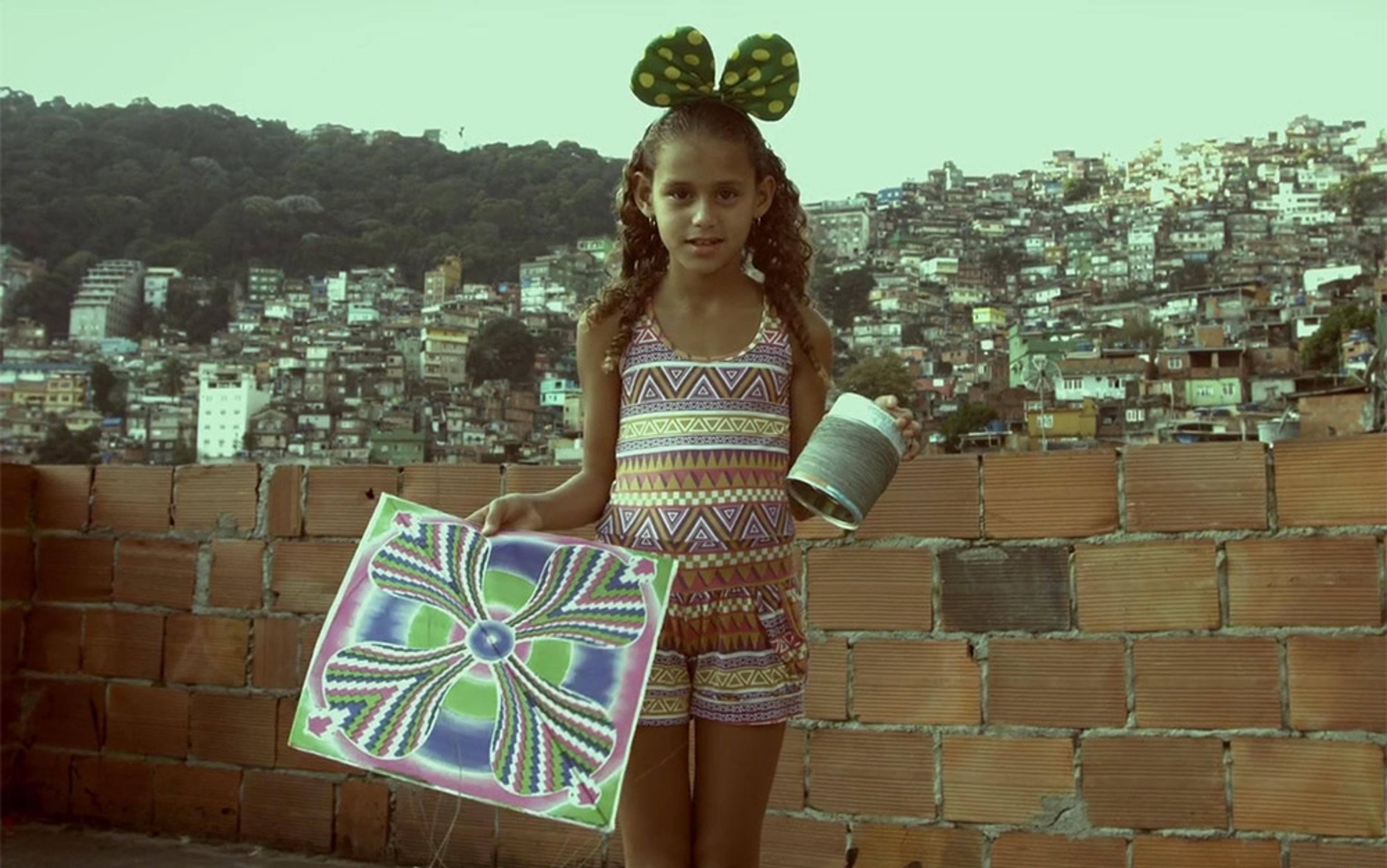 A girl holding a kite and string spool with a cityscape and hillside in the background, standing by a brick wall.