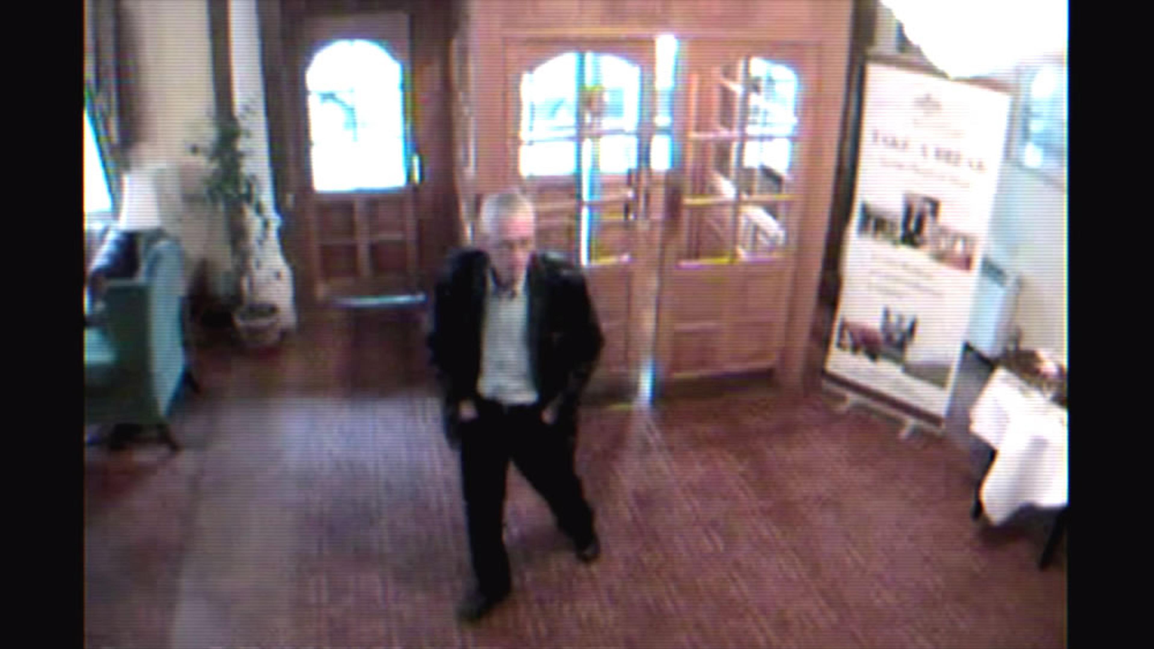 A CCTV image showing a man walking in a lobby area with double wooden doors, a signboard, a plant and seating visible.