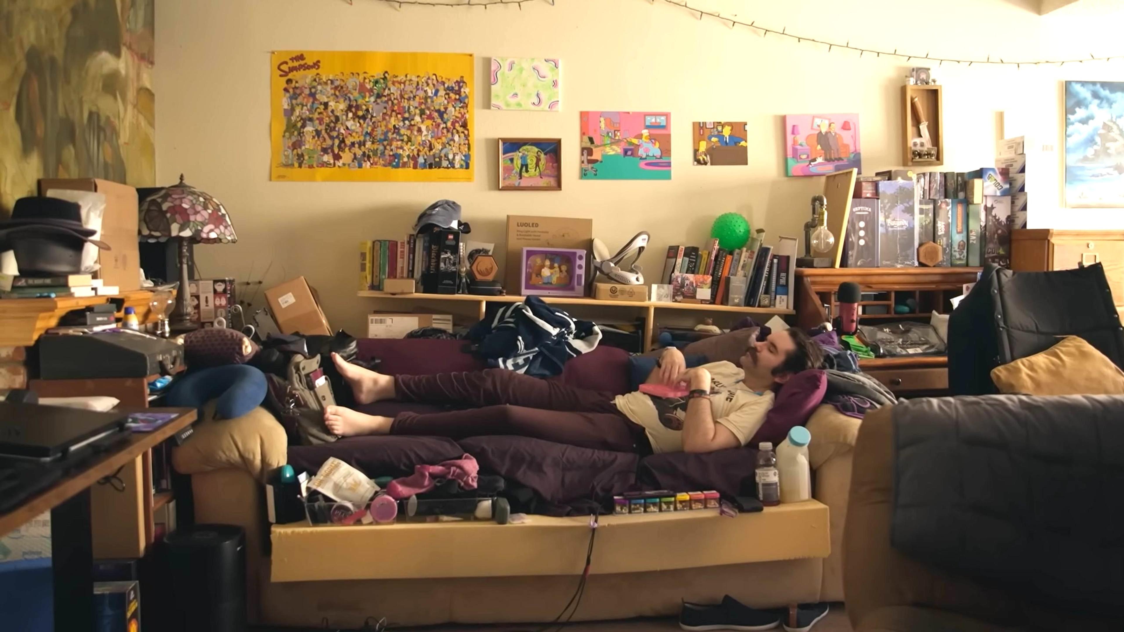 Photo of a messy room with a person lying on a cluttered sofa surrounded by books posters boxes and string lights.