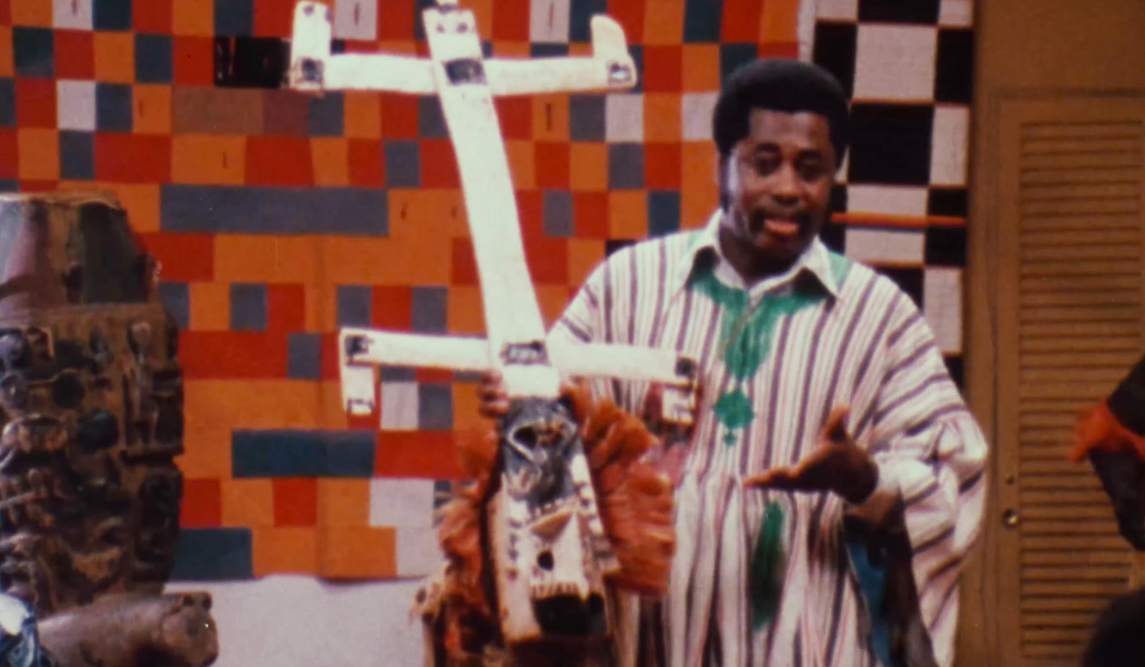 A man in a striped shirt holds a white mask, with a red, grey, and black patterned background.