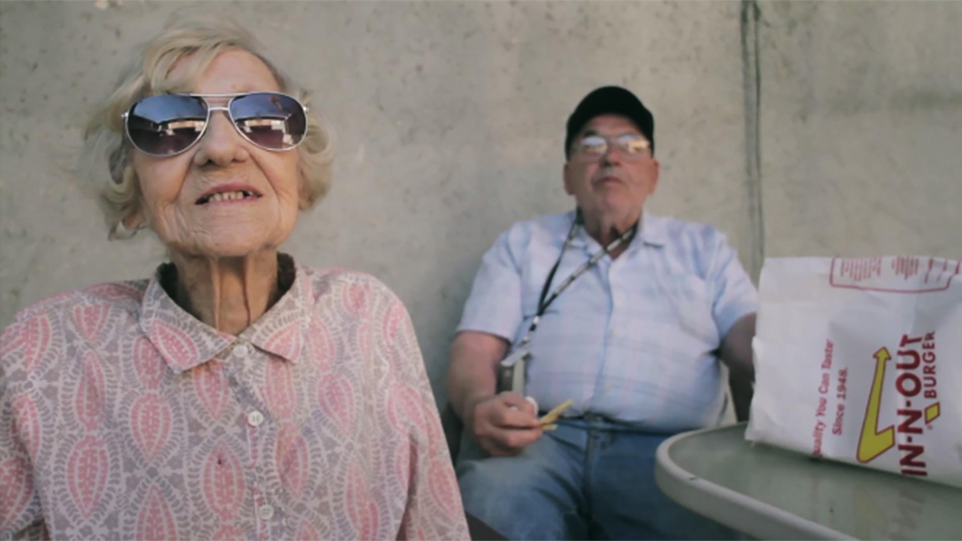 An elderly woman in mirrored sunglasses and a pink blouse, and an elderly man eating fries next to her with a takeout bag on the table.