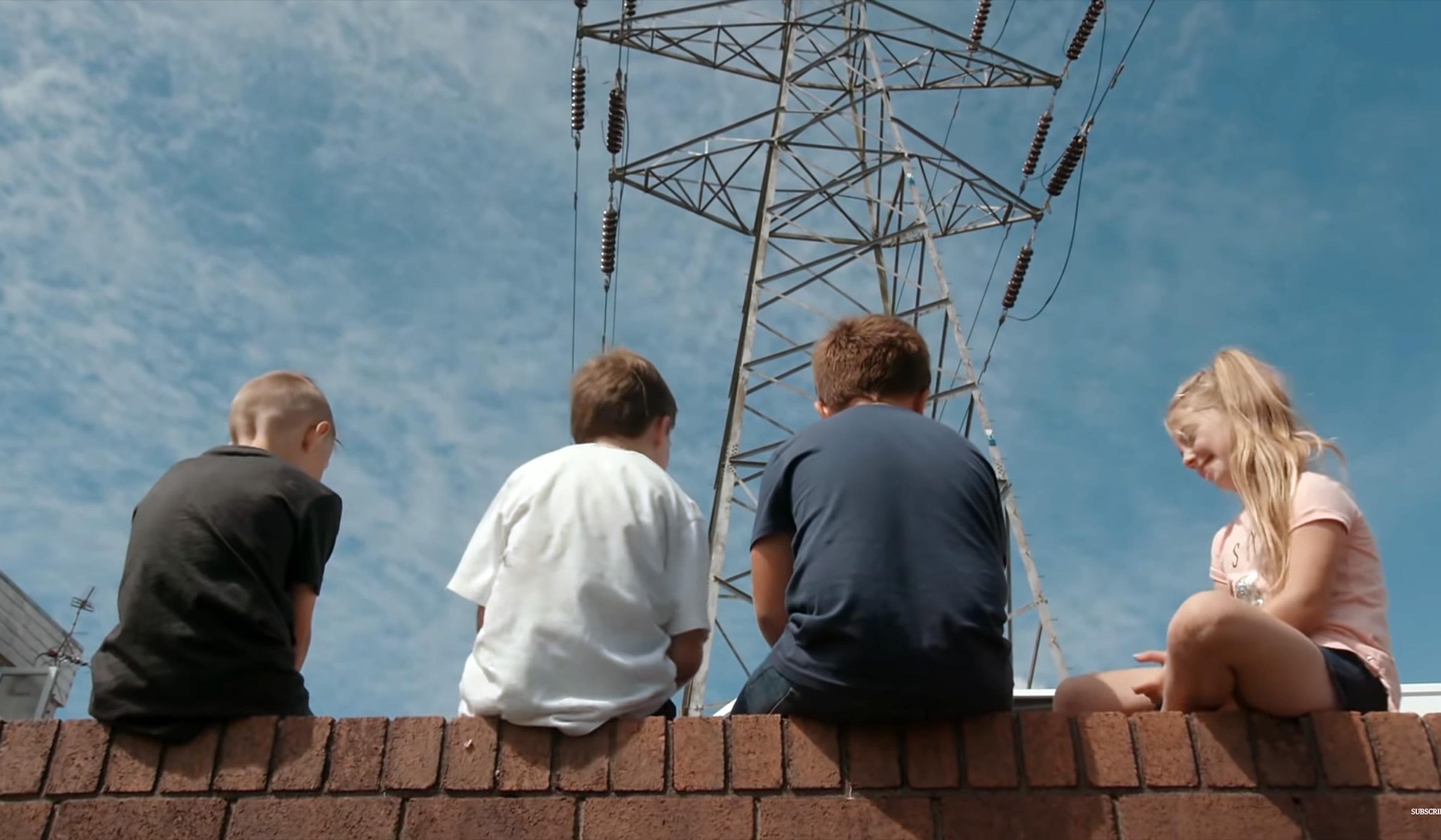 Four children sitting on a brick wall with their backs to the camera, an electricity pylon visible behind them against a blue sky.