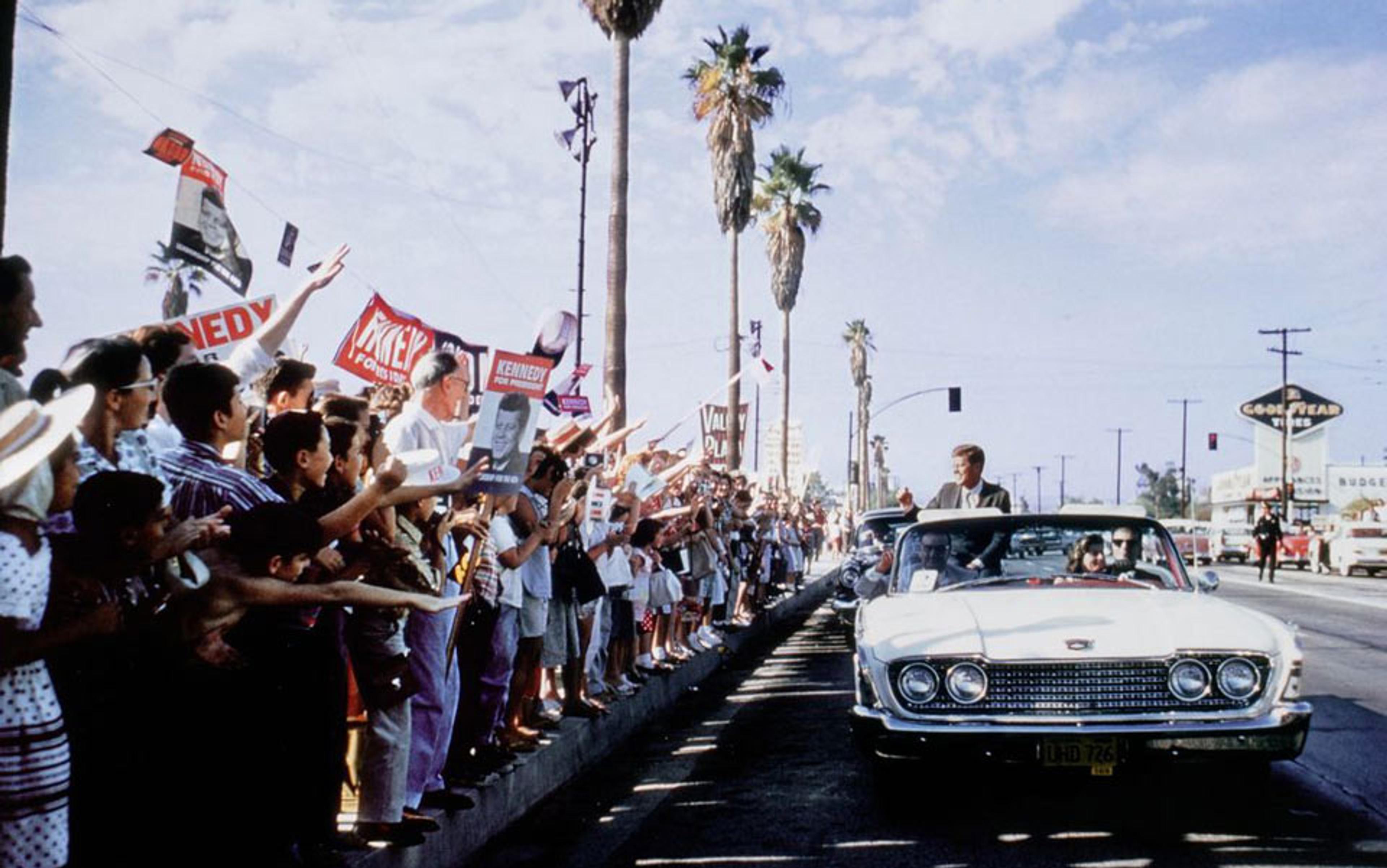 President John F Kennedy in a white convertible waving to a crowd holding “Kennedy” signs during a parade on a street lined with palm trees.