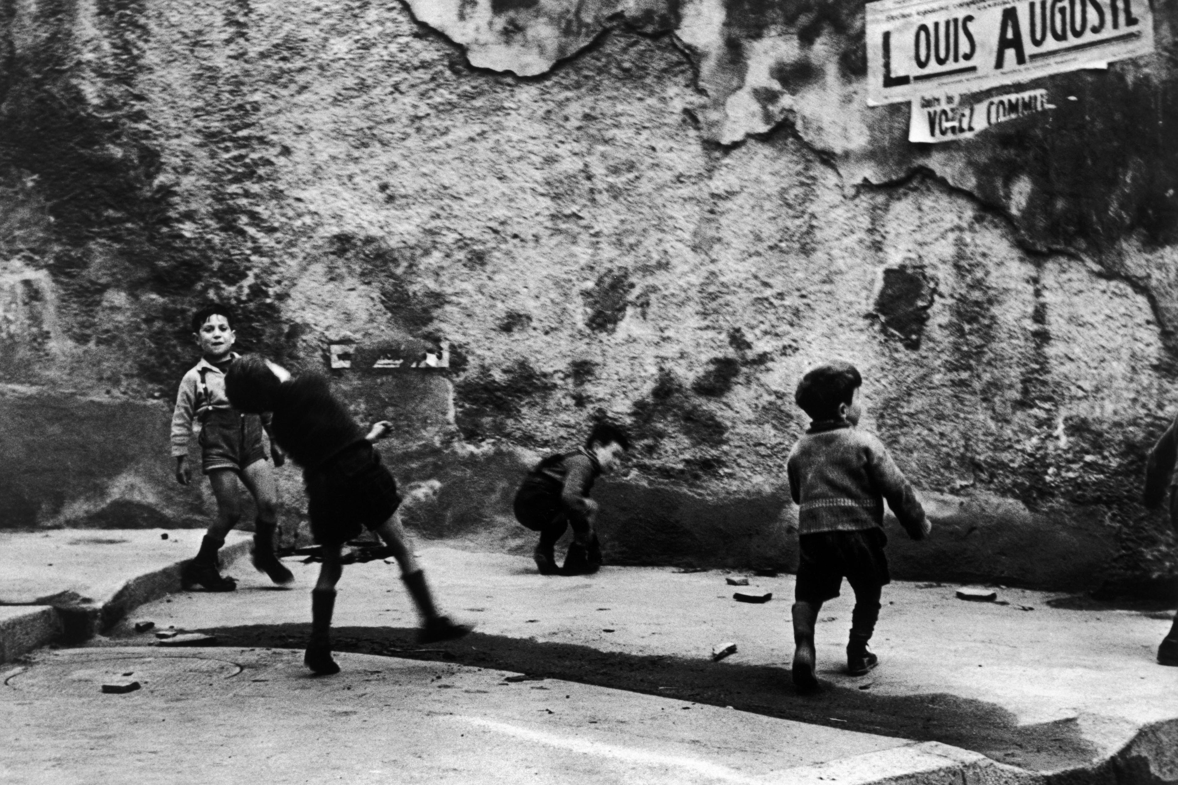 Black and white photo of children playing in a street against a rough wall with a sign, some moving energetically.
