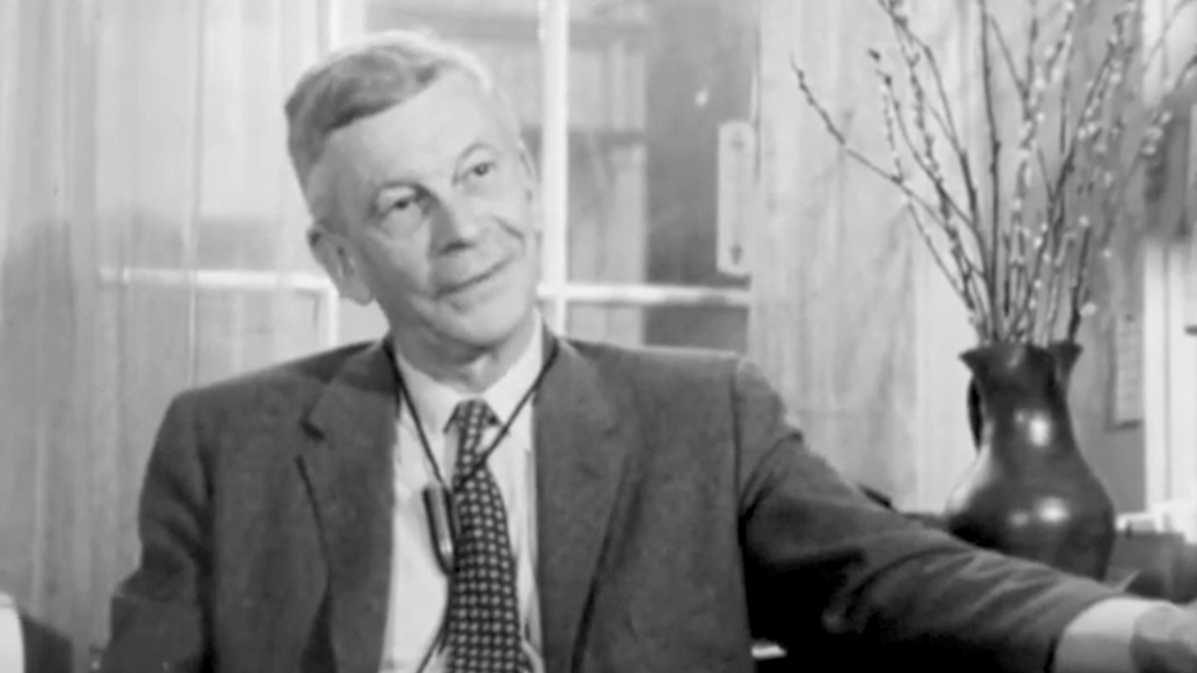 Black and white photo of a man in a suit sitting indoors with a vase and branches.