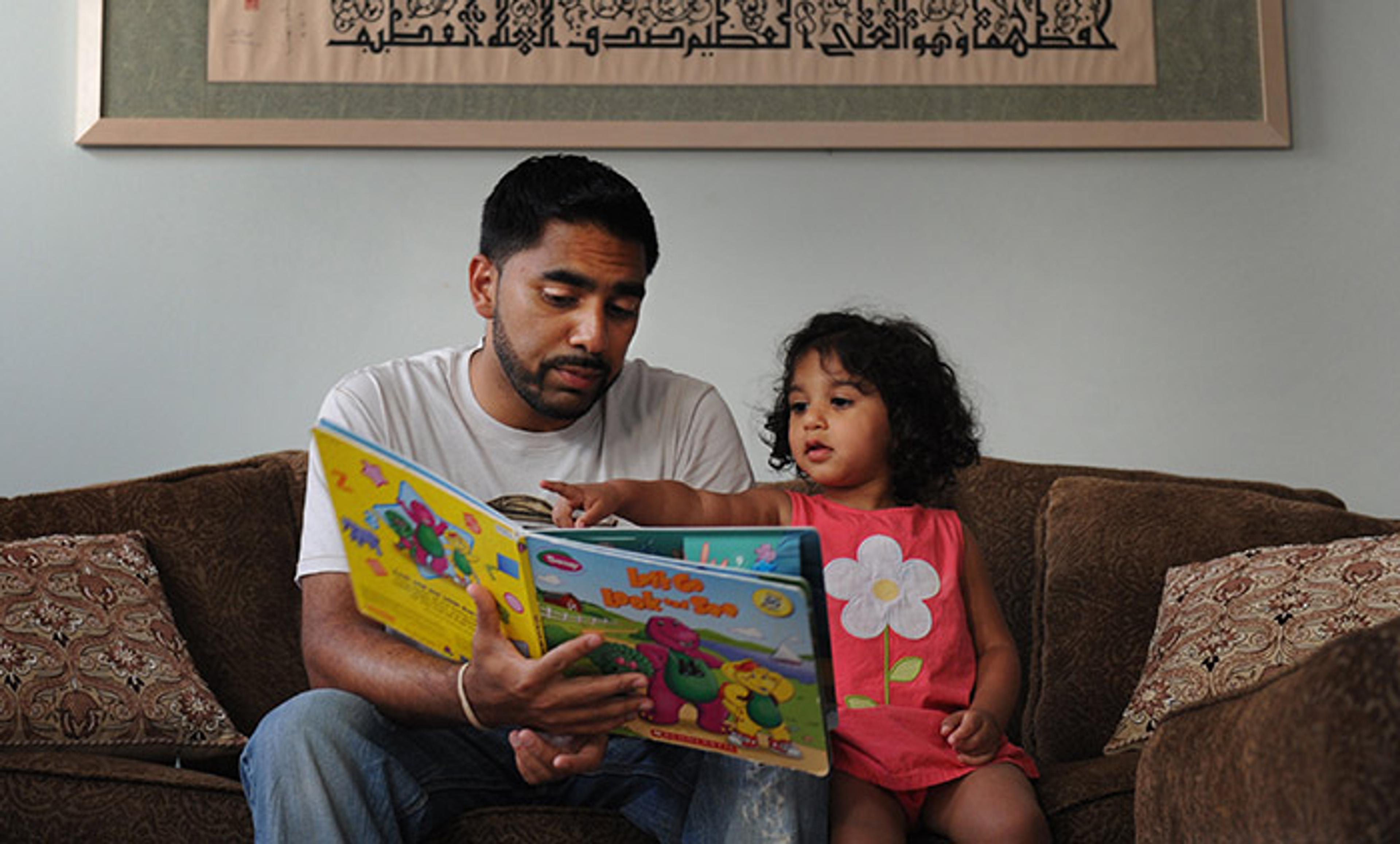 An Arab man adult reading a colourful Western picture book to a girl on a sofa with a framed artwork featuring Arabic calligraphy on the wall.