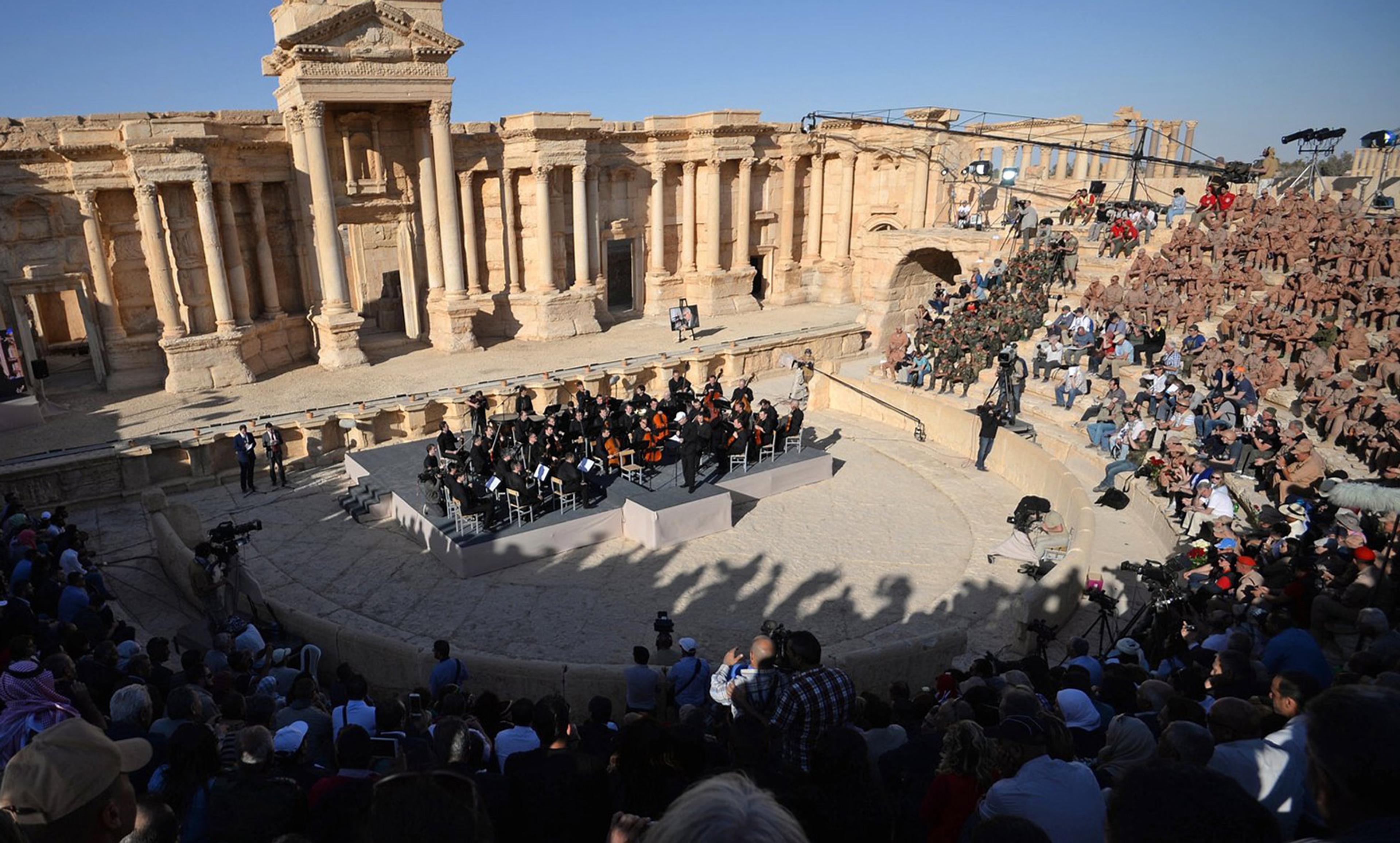 An outdoor orchestra performance at an ancient amphitheatre with a large audience and historic stone architecture.