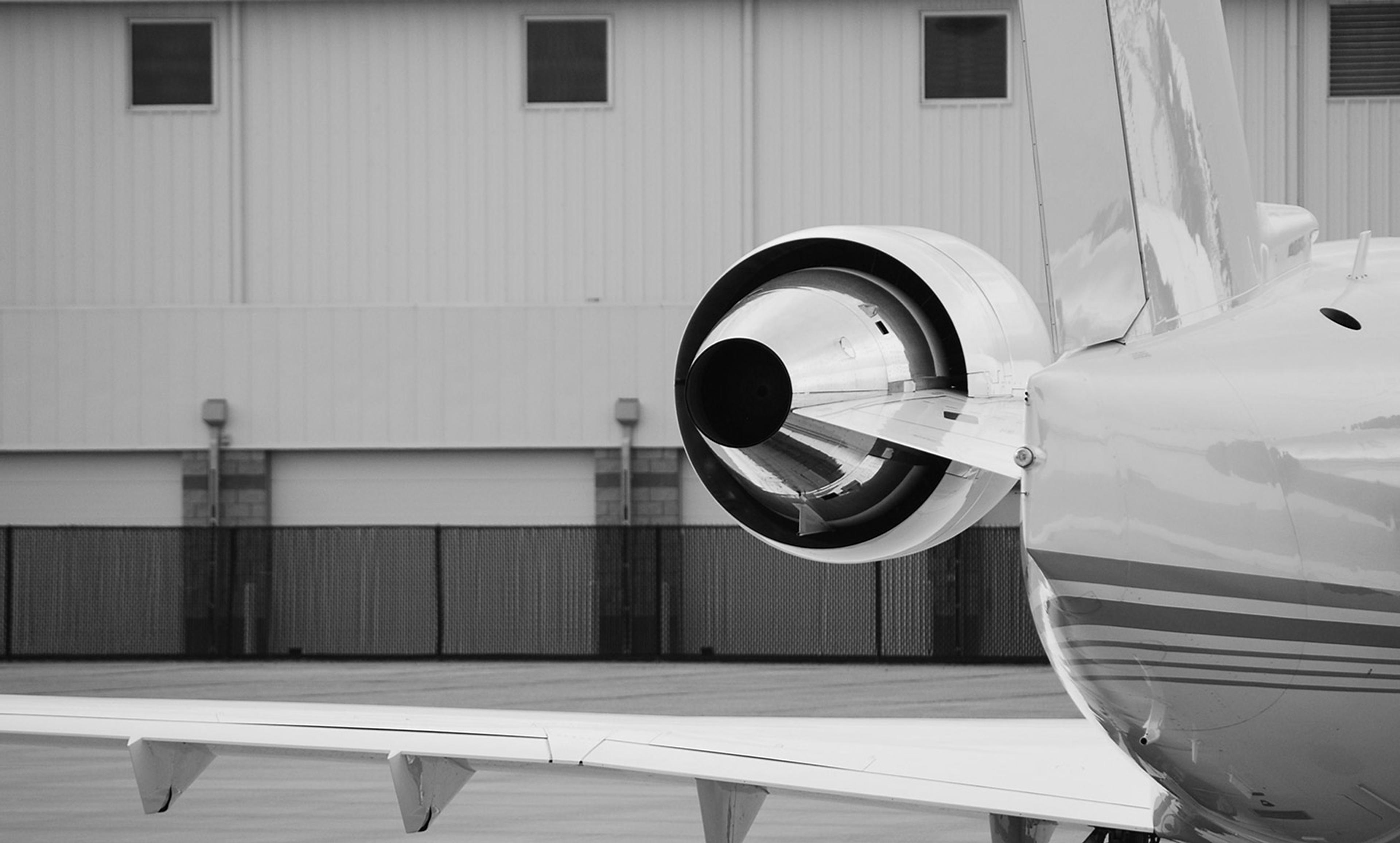 Black and white photo of a jet engine and its rear section on the tarmac, with a hangar wall in the background.