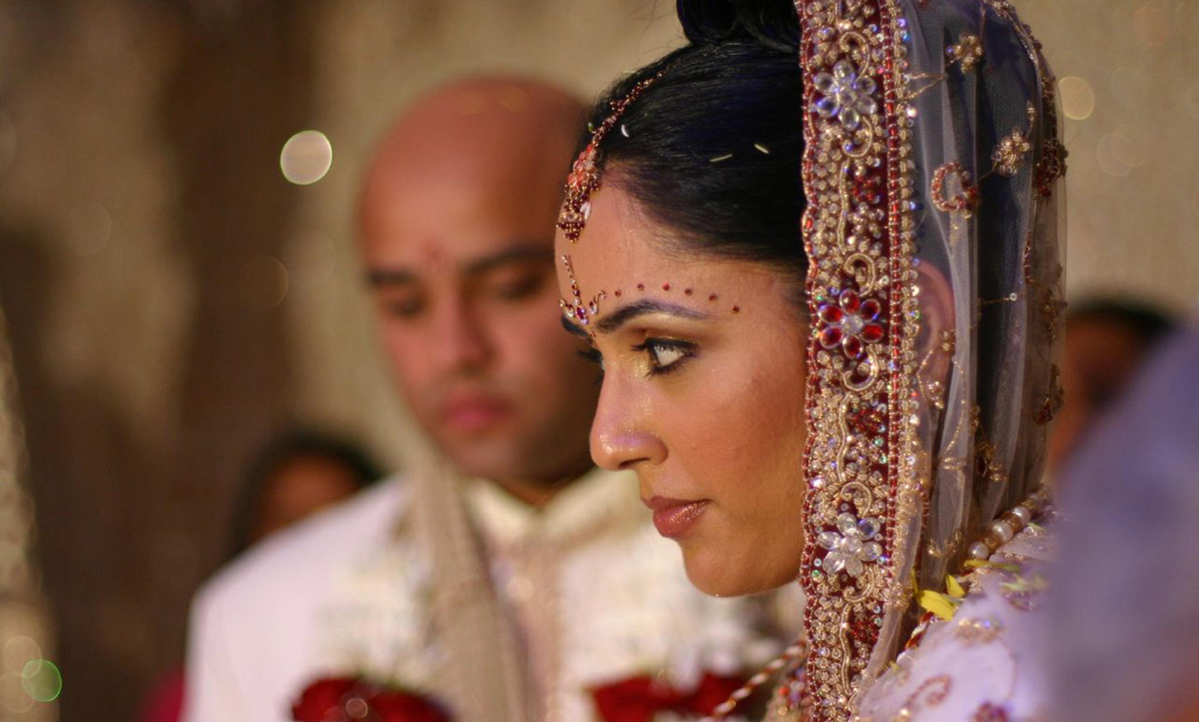 A bride in traditional Indian attire with a groom in the background, both adorned in intricate jewellery during a wedding ceremony.