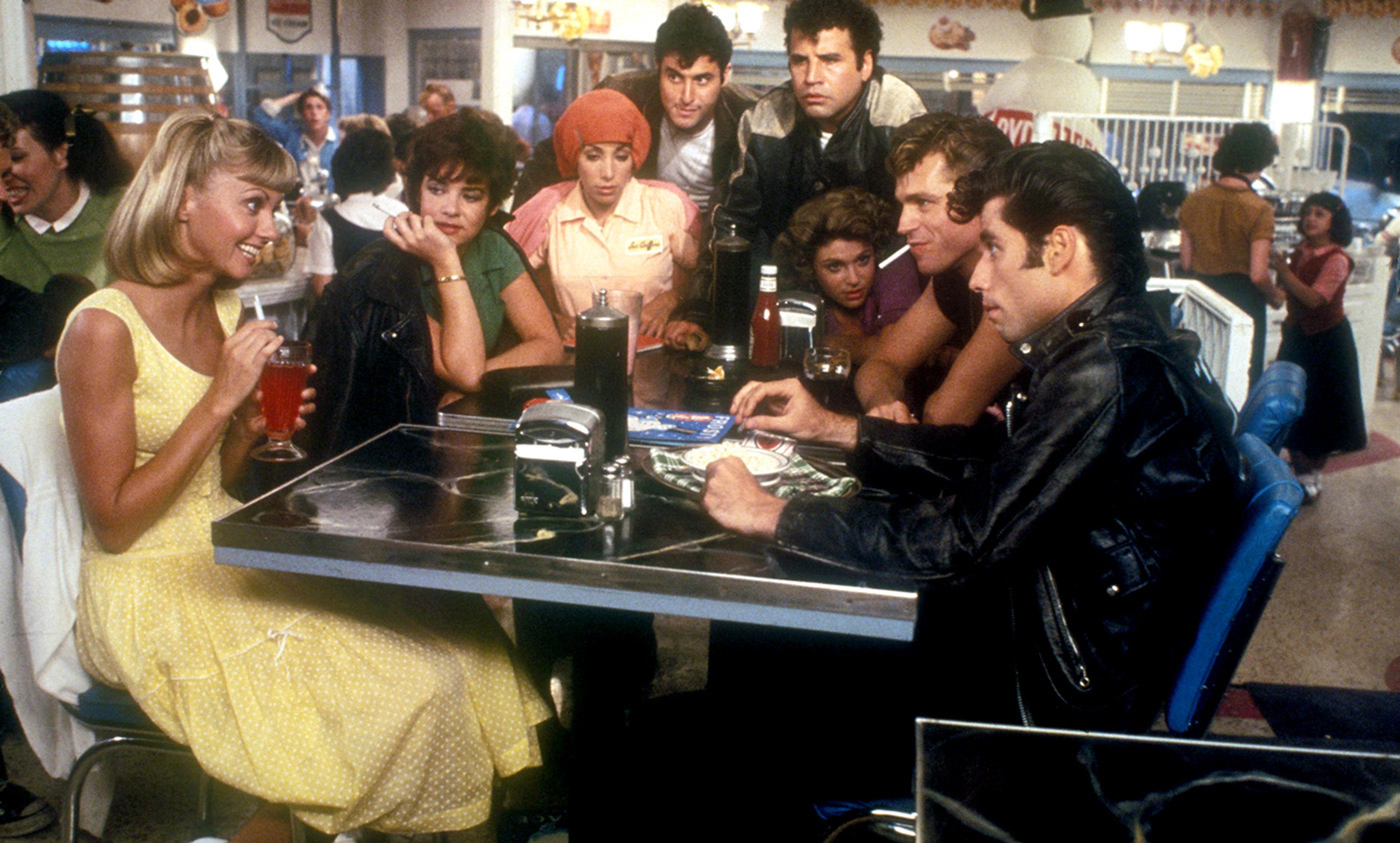 Film still from Grease showing a young crowd in 1950s-style diner, with Olivia Newton John in a yellow dress smiling at John Travolta in a booth.