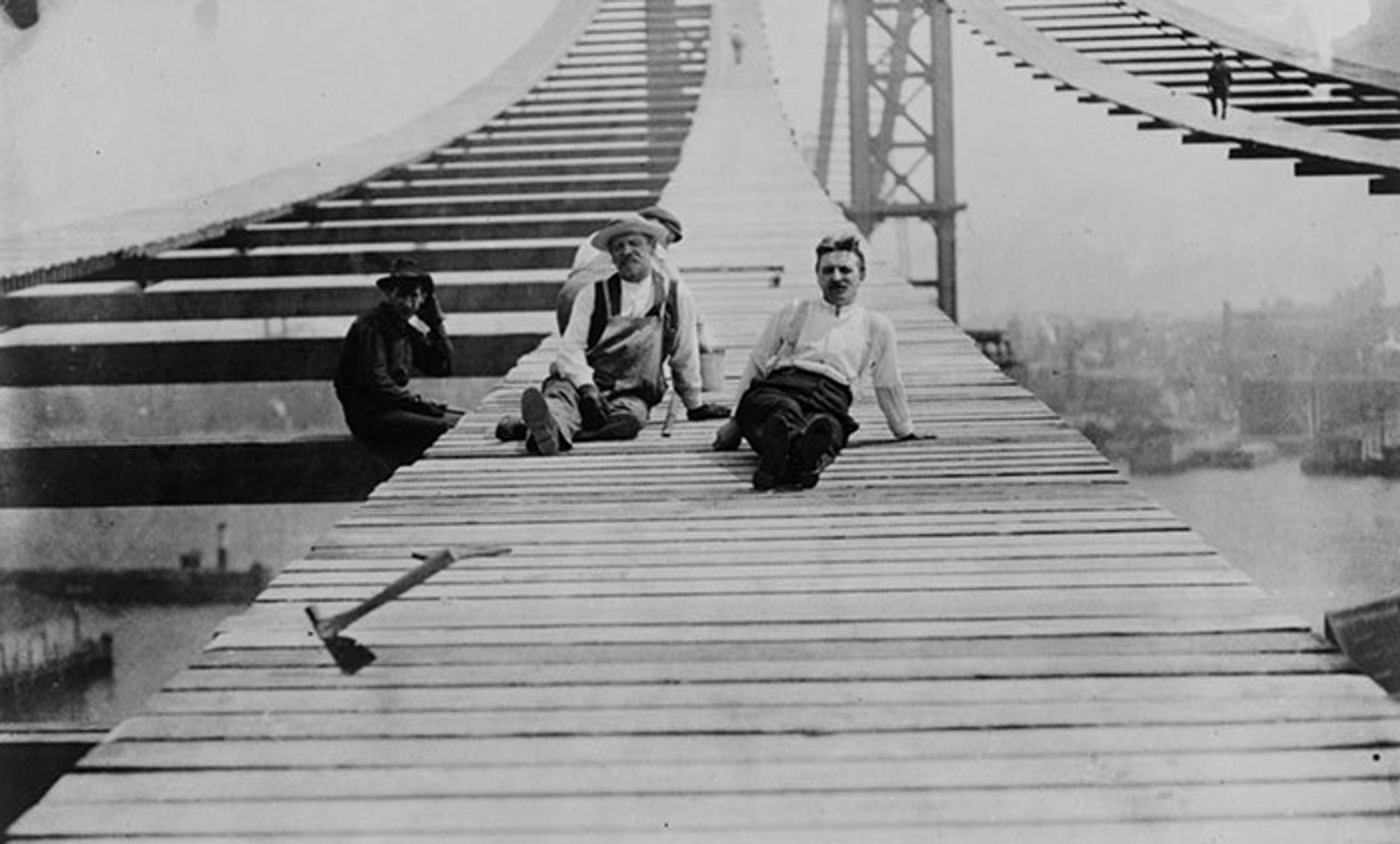 Three men sitting on the incomplete deck of a suspension bridge under construction, with cityscape in the background.