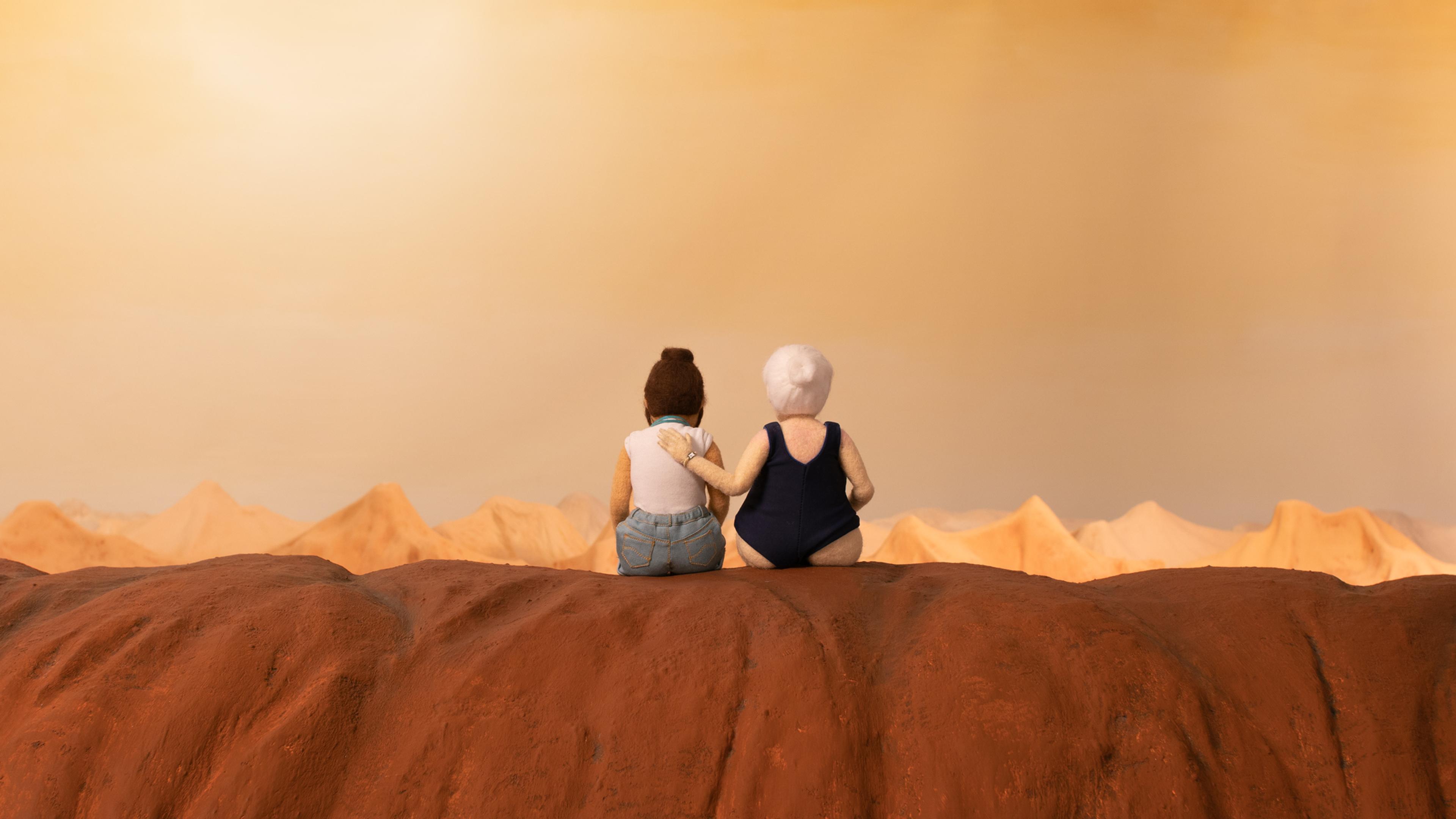 Two felt figures sit together facing a desert landscape under a warm sky.