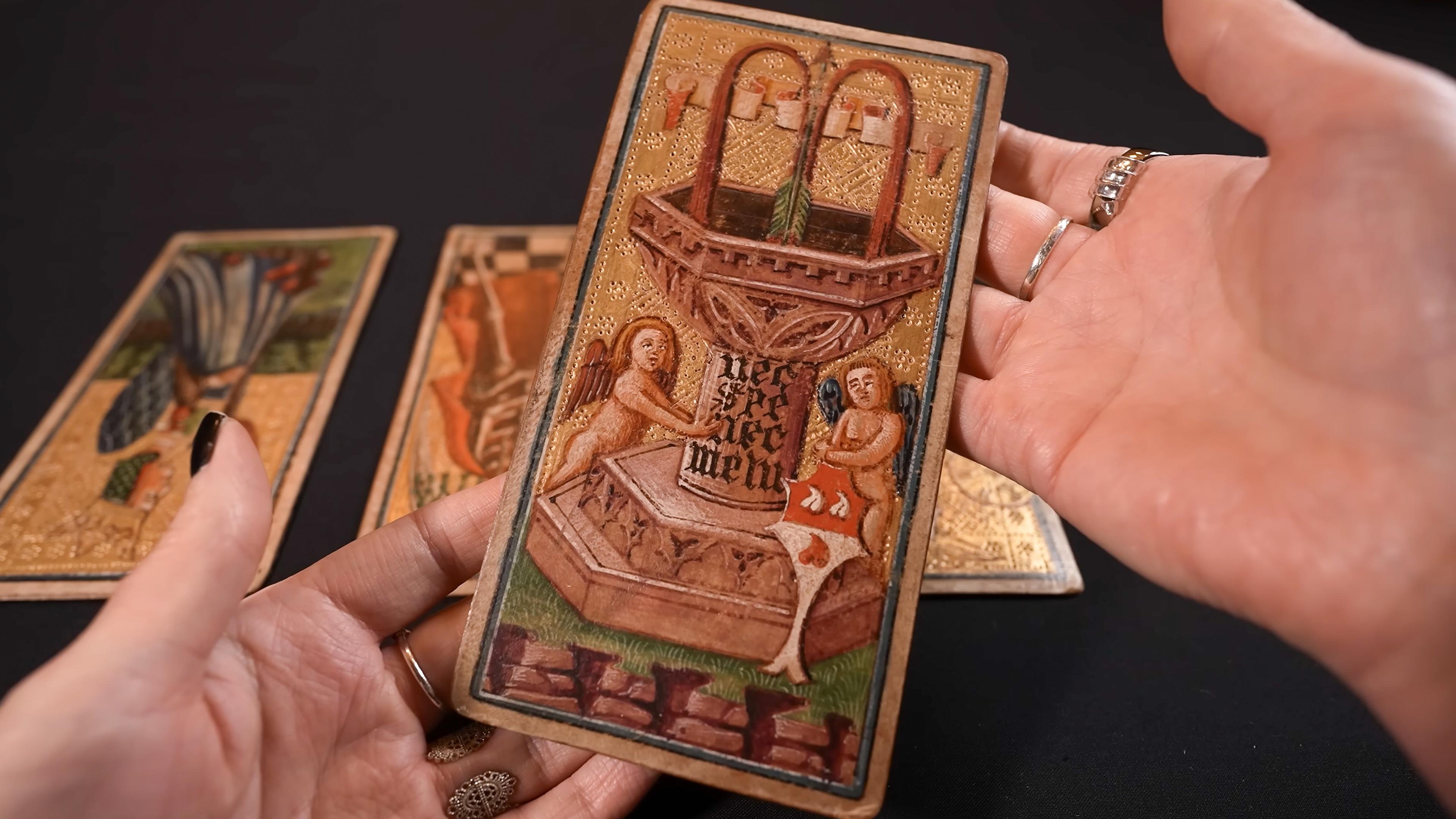 Photo of a hand holding an ornate tarot card with a decorative illustration, two other cards on a black surface in the background.