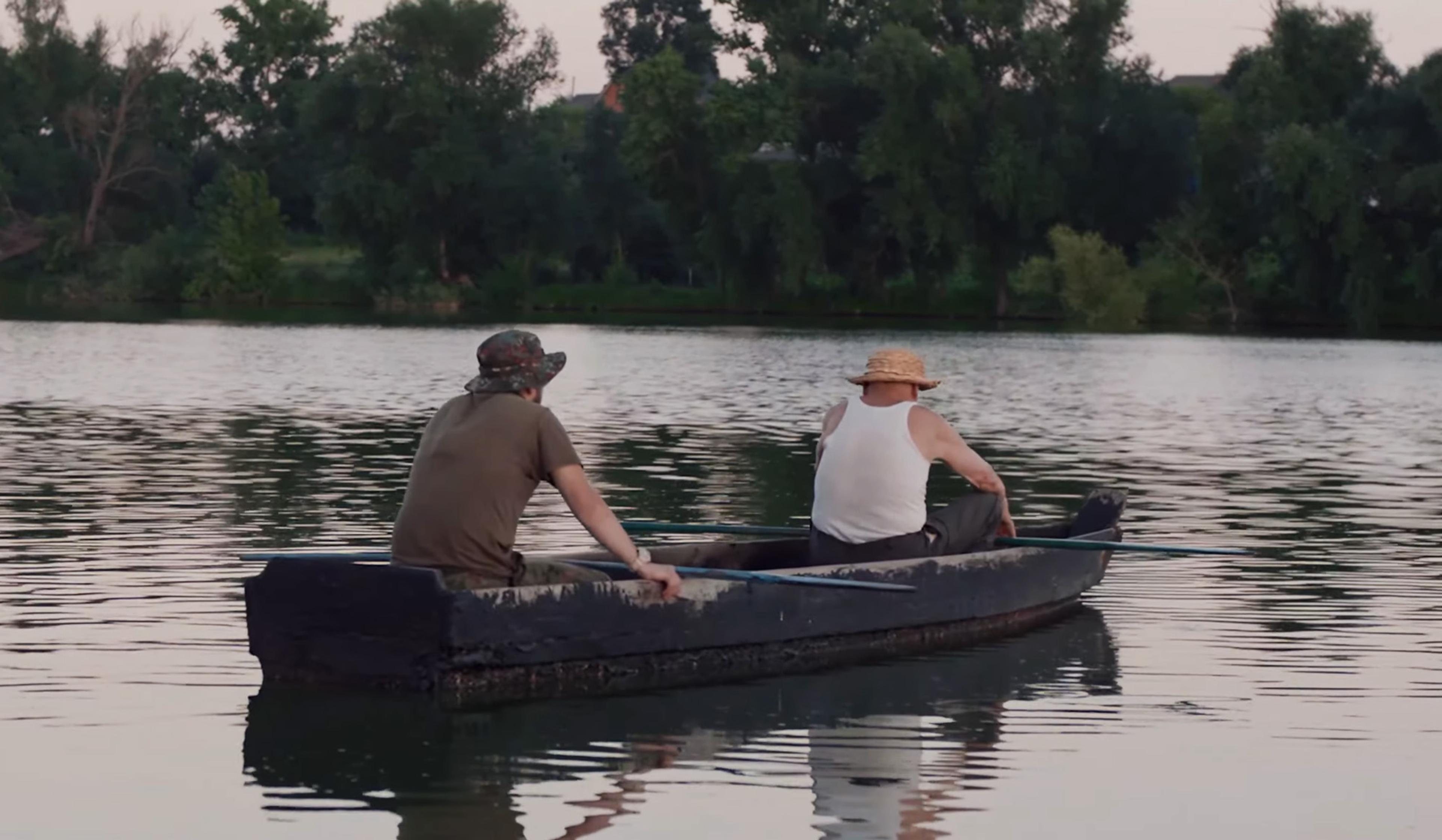 Two men wearing hats are in a small boat on a calm lake, surrounded by trees. One is rowing while the other looks on.