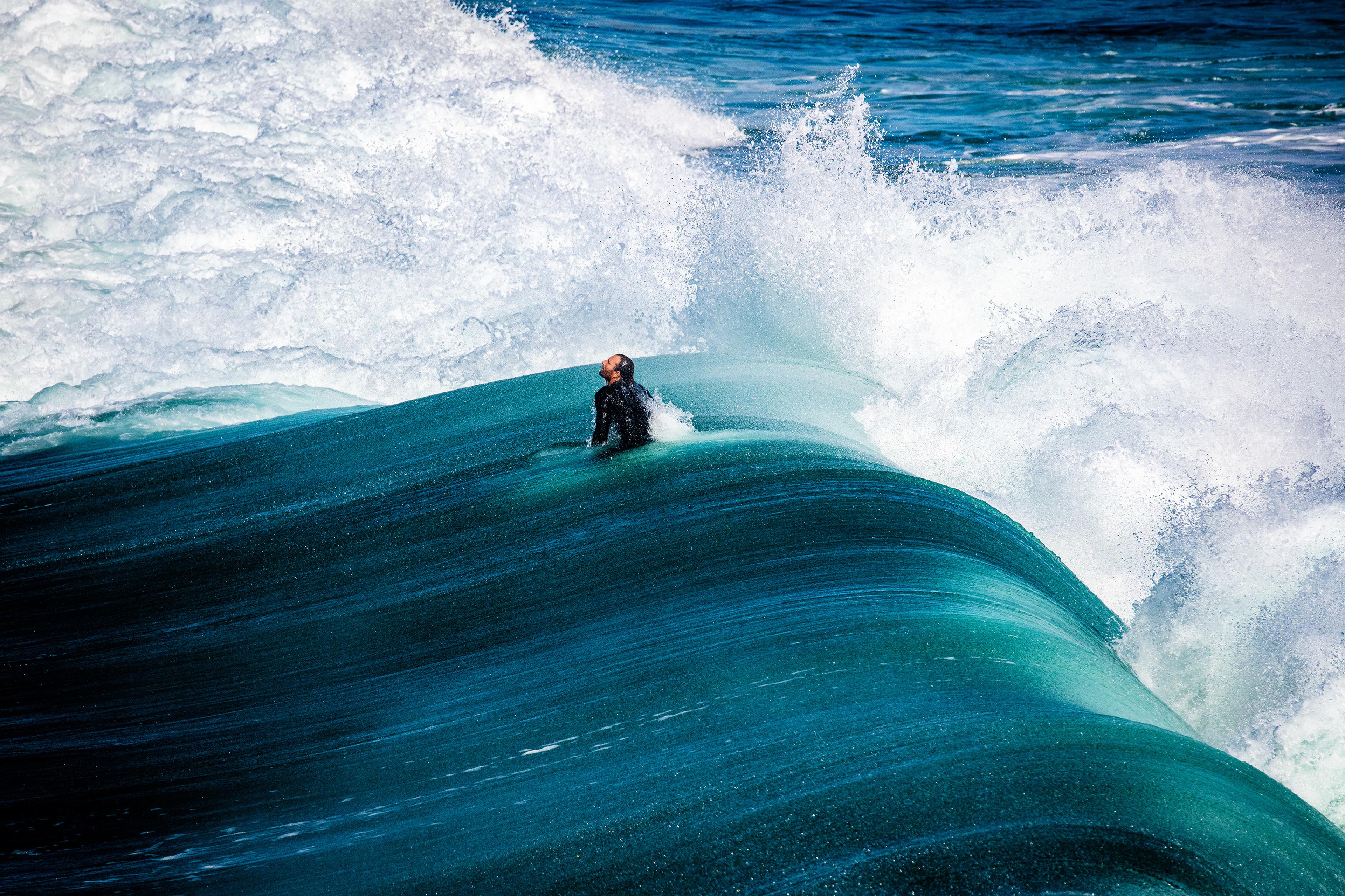 A surfer in a wetsuit emerging from a large turquoise wave with white foam crashing around.