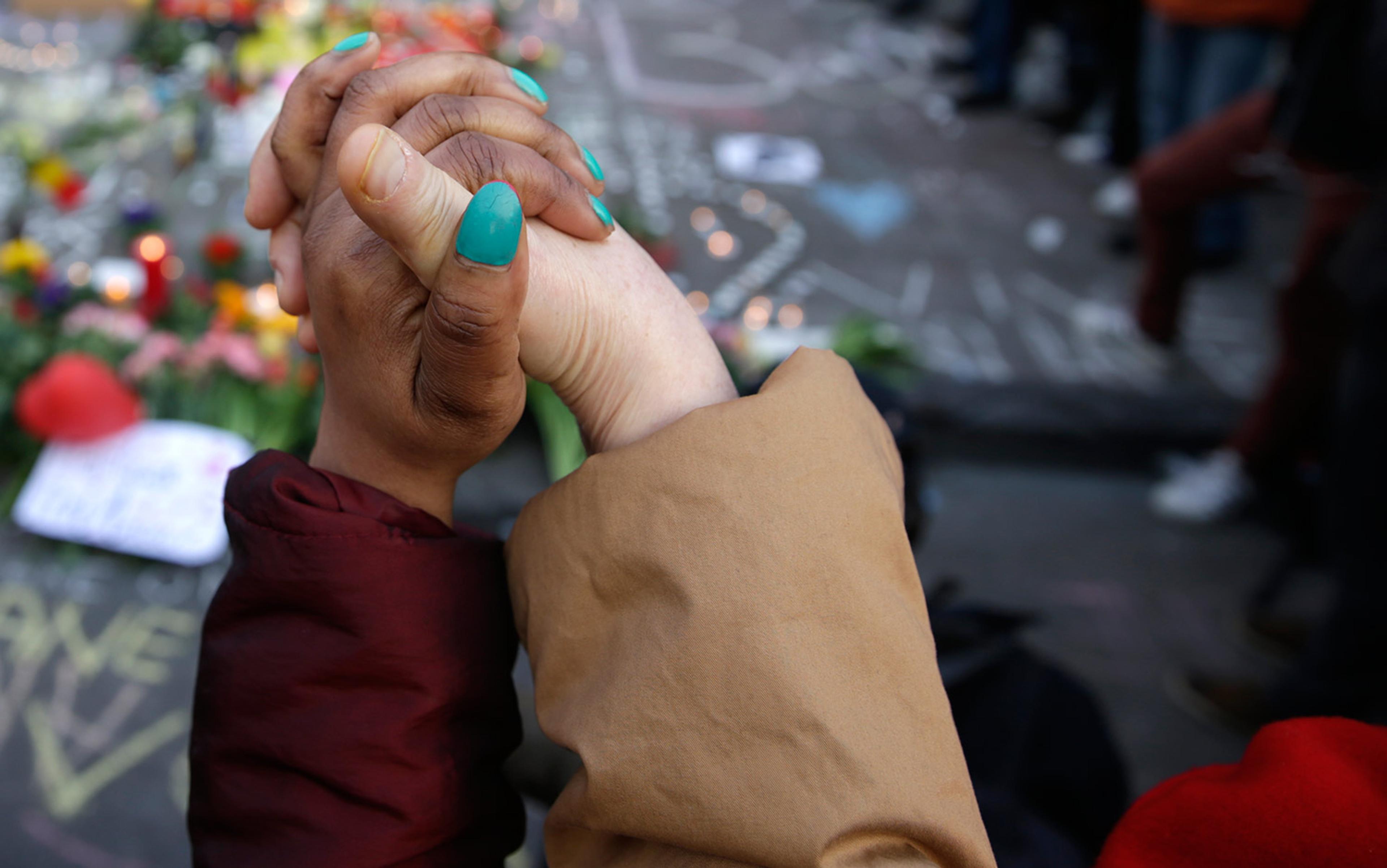 Photo of two hands clasped tightly, one lighter-skinned and one darker-skinned, with a background of a memorial with flowers and candles.