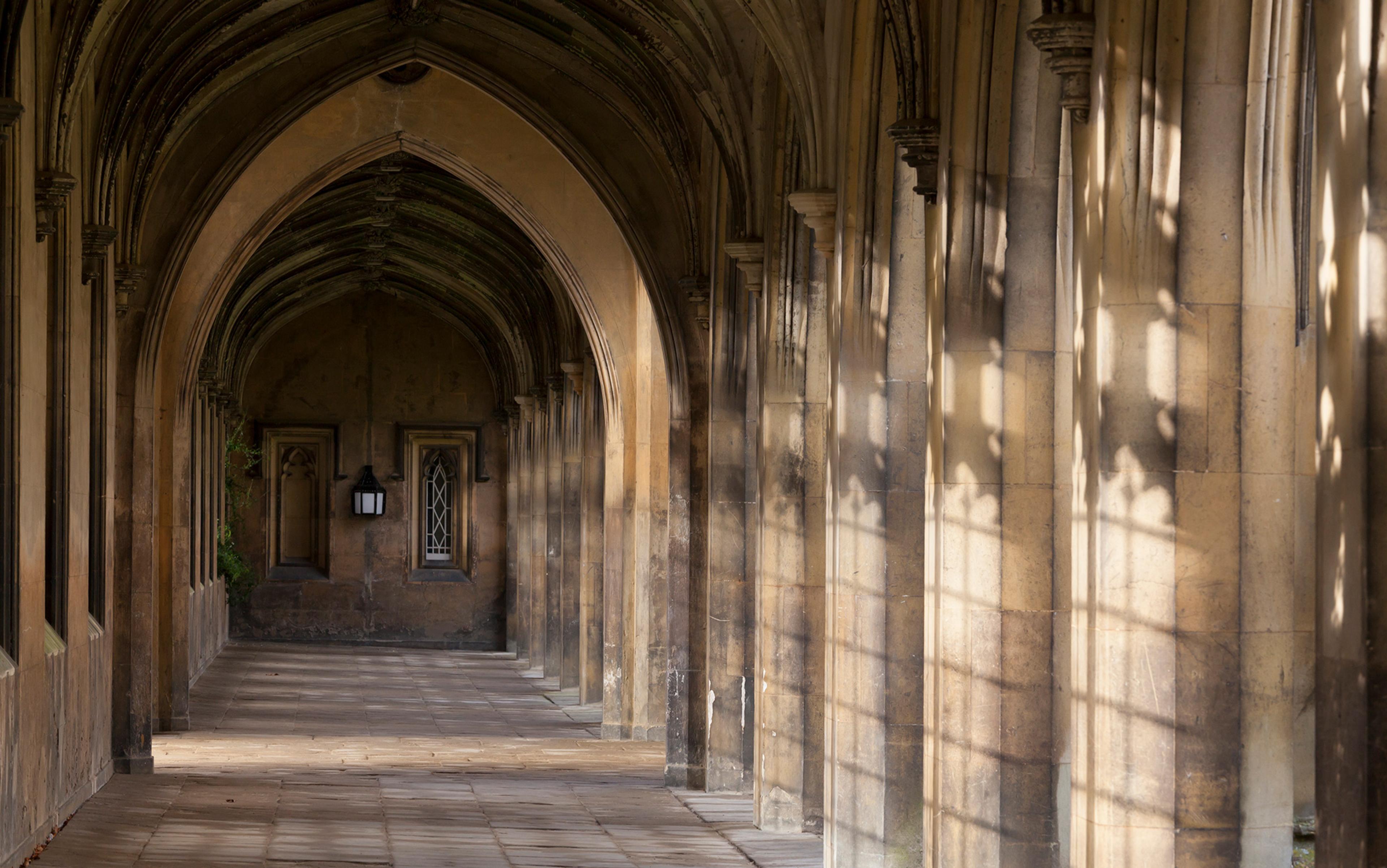 A sunlit old stone corridor with Gothic arches and shadows cast on the walls, featuring arched windows and a lantern.