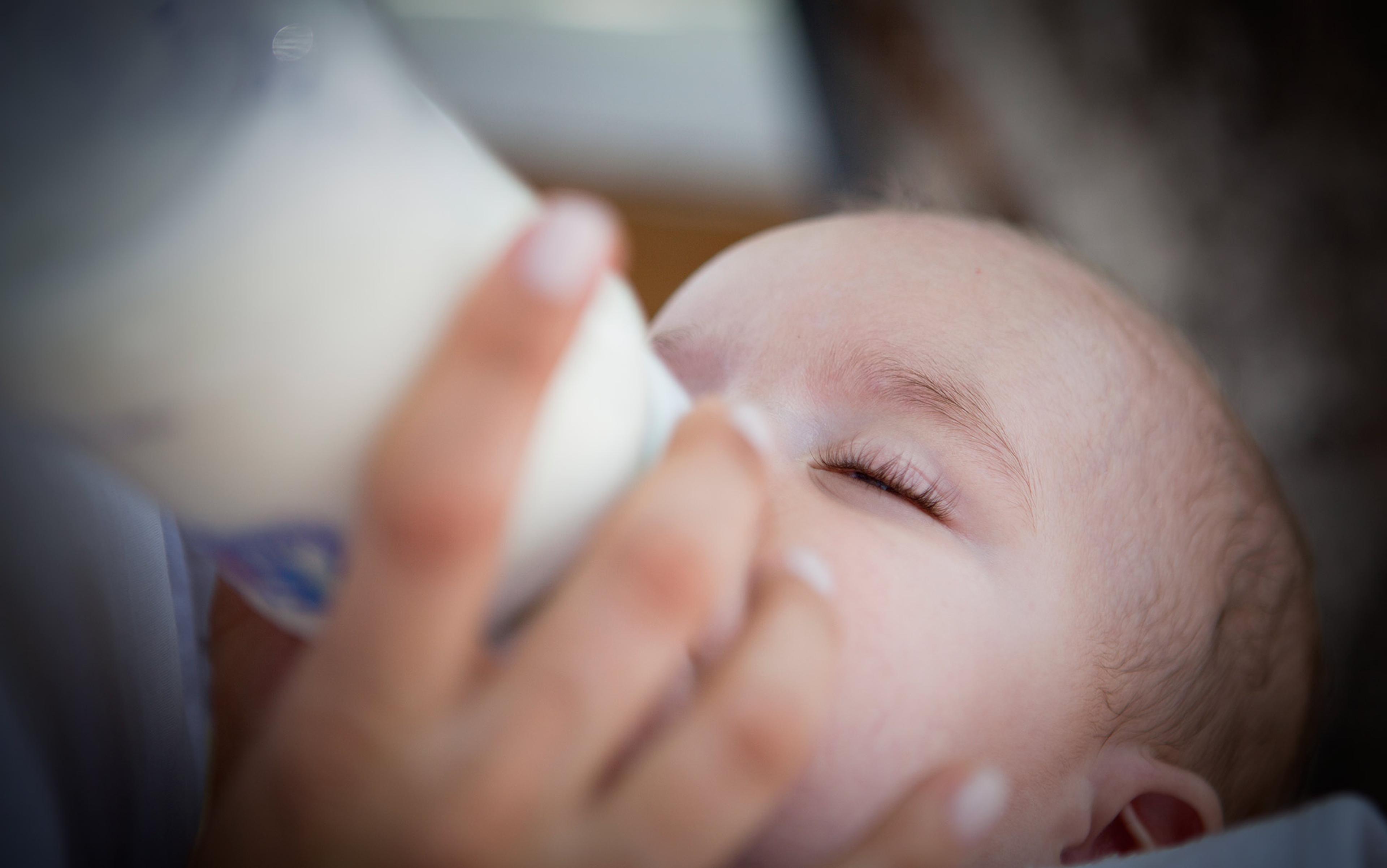 A baby drinking milk from a bottle, eyes closed with a relaxed expression, held closely by an unseen person.