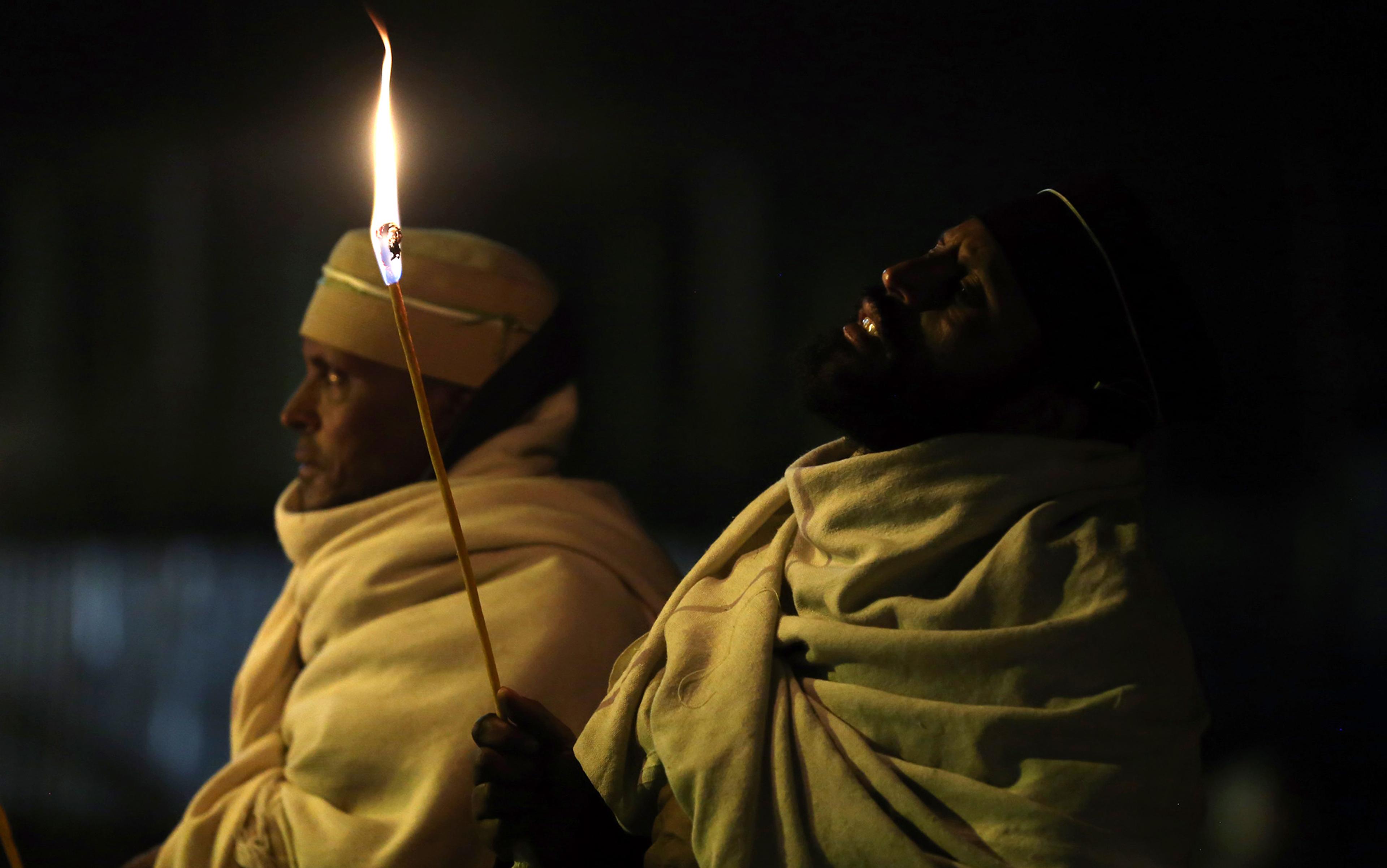Two people wearing light-coloured ceremonial robes holding lit candles in a dark setting. One looks ahead, the other slightly tilts up.