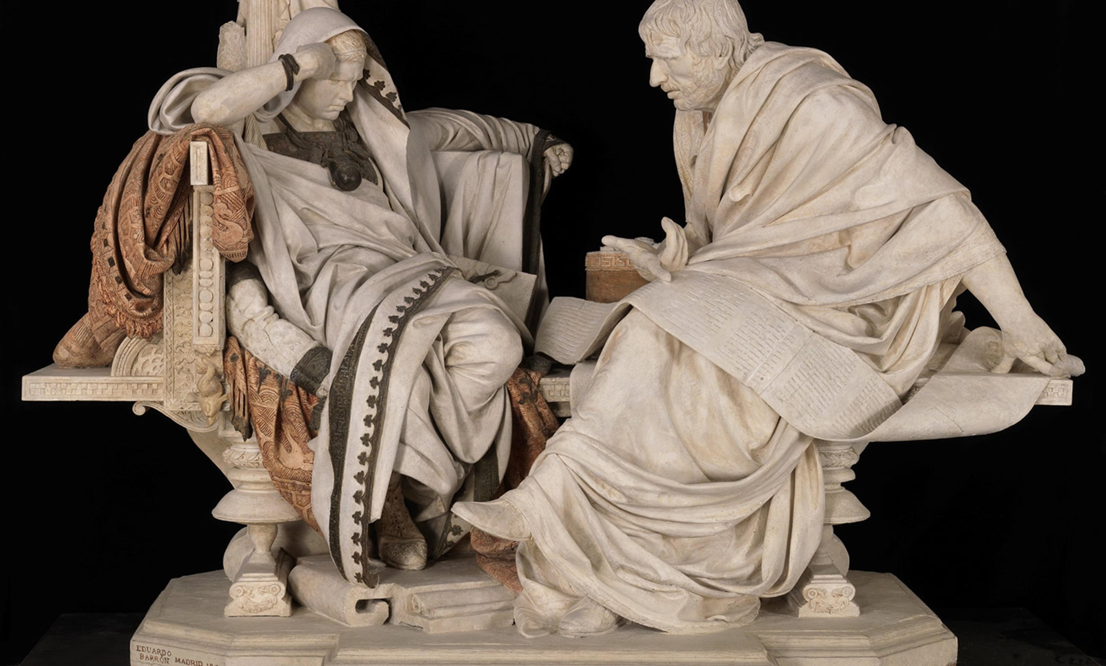 Marble statue depicting two historical figures in discussion, one sitting with a scroll. The statue is detailed with draped robes.