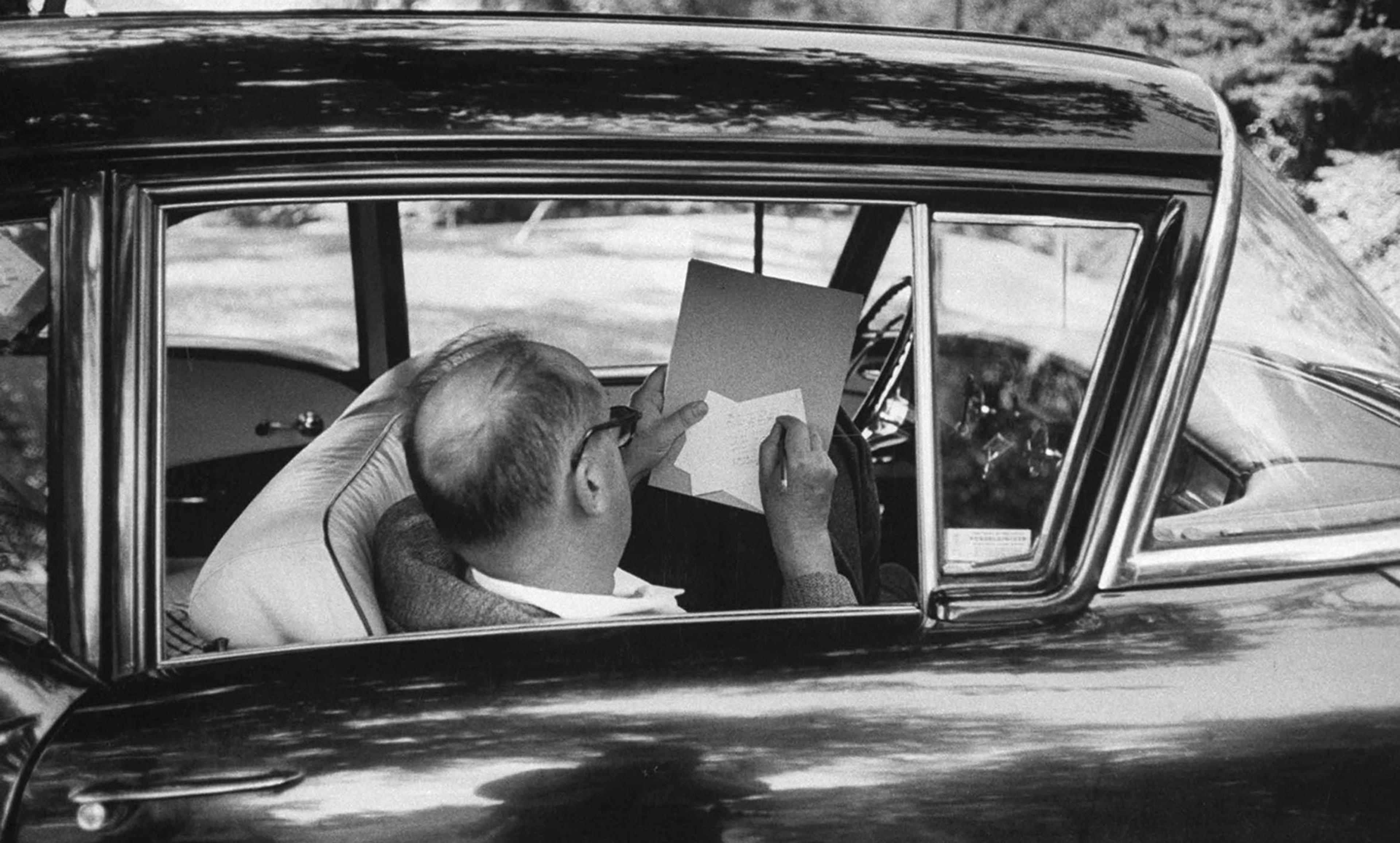 Vintage-looking Black and white photo of a man in a car viewed from the rear side window, writing on paper with a pencil.