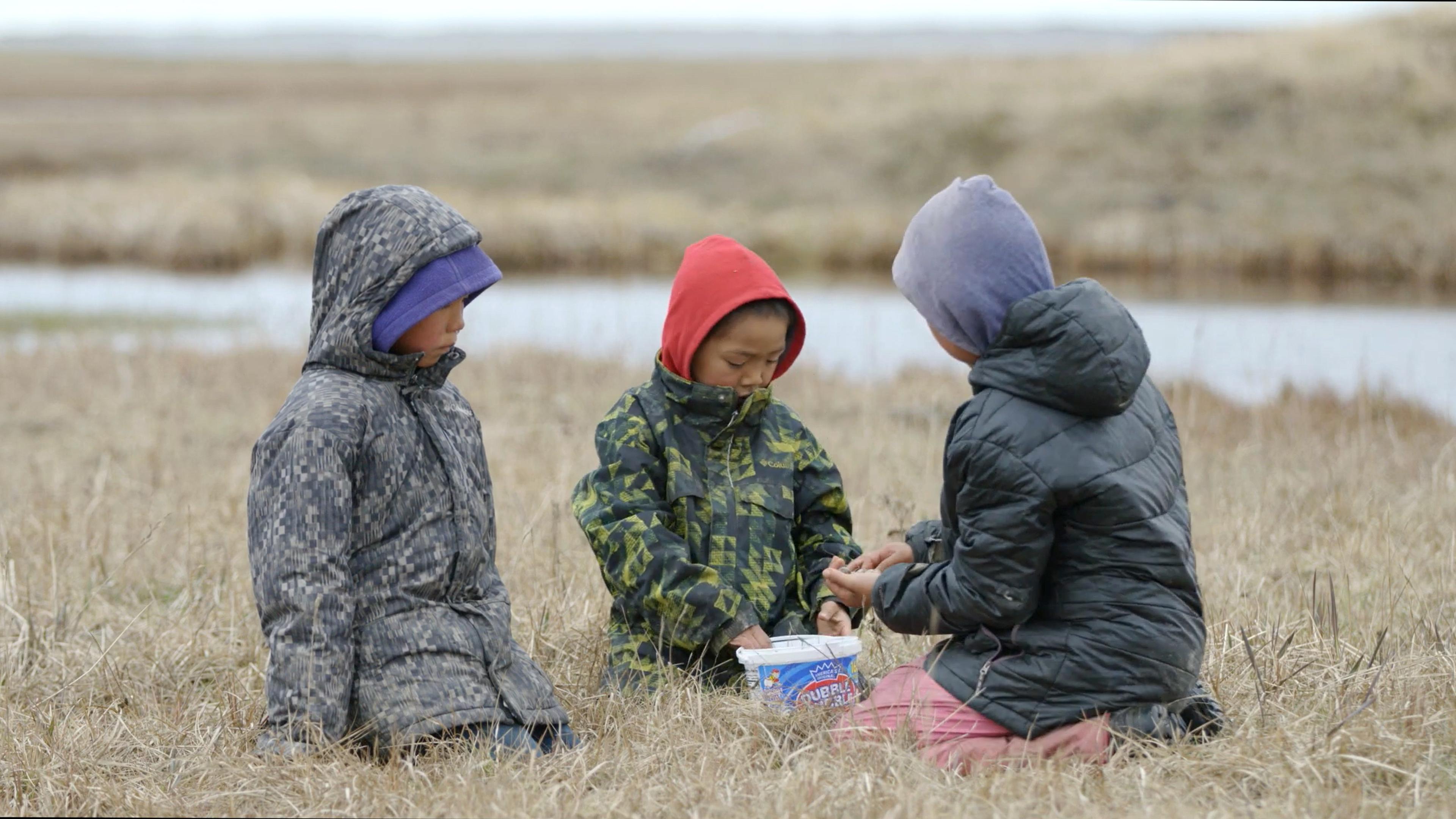 Photo of three children in winter jackets sitting on grassland near a stream with a plastic container between them.