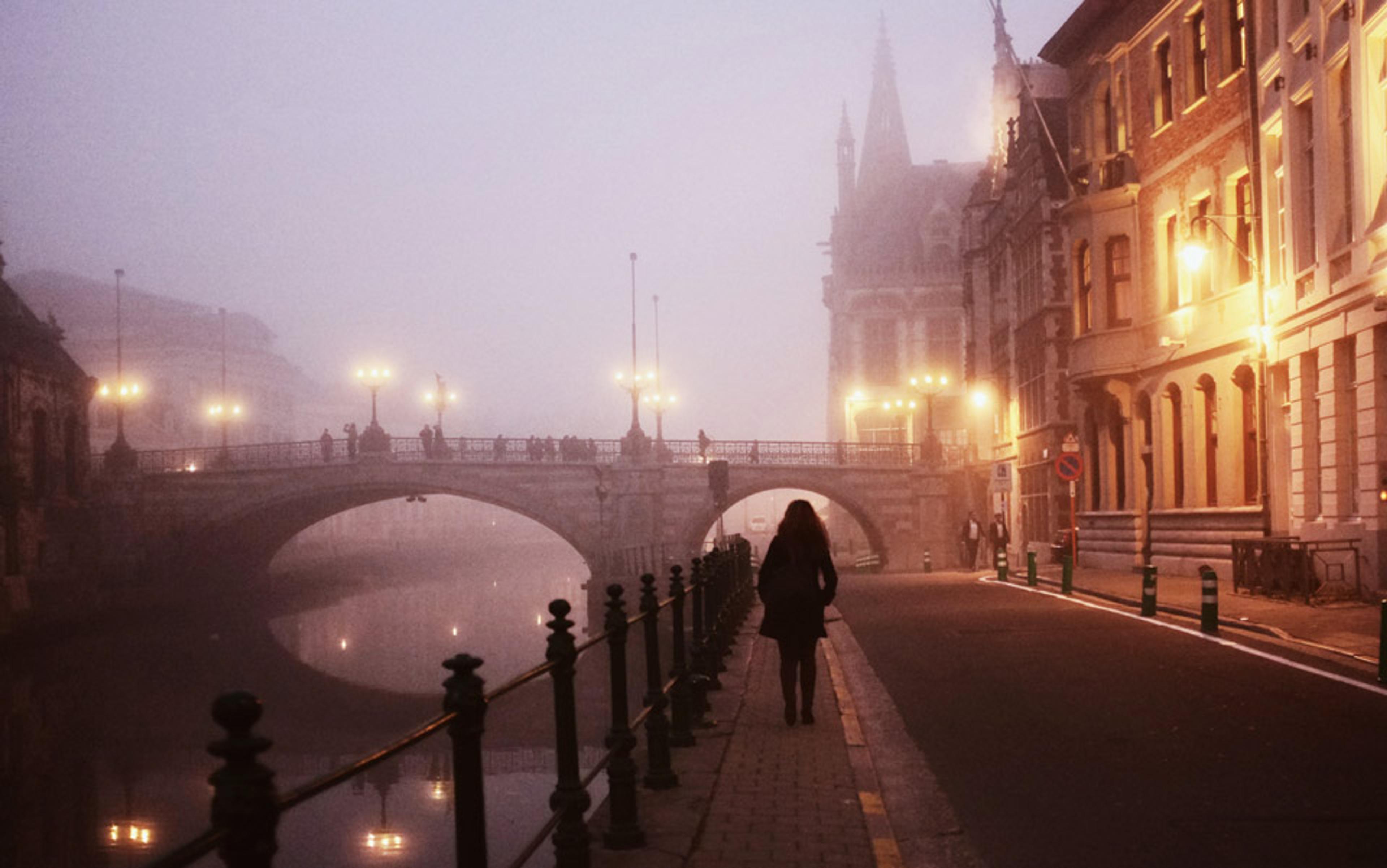A person walking along a foggy street by a river, with a lit bridge and historical buildings in the background.
